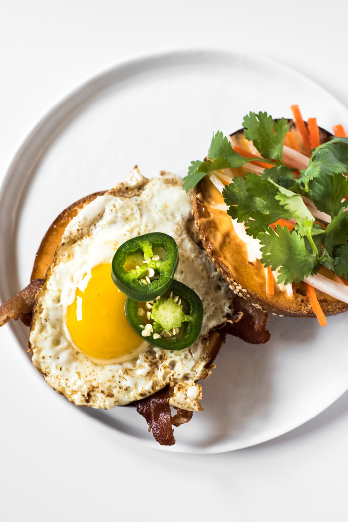 This Breakfast Bánh Mì is a bagel topped with bacon, a fried egg, Sriracha-mayo, do chua, sliced jalapeno, and cilantro. Bring lots of napkins!